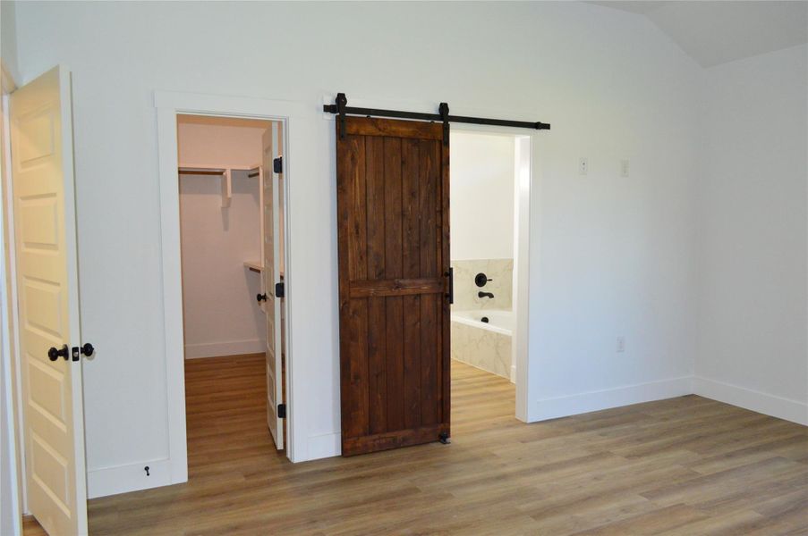 Unfurnished bedroom with a barn door, light wood-style flooring, ensuite bath, and a walk in closet Unfurnished bedroom with a barn door, light wood-style flooring, ensuite bath, and a walk in closet
