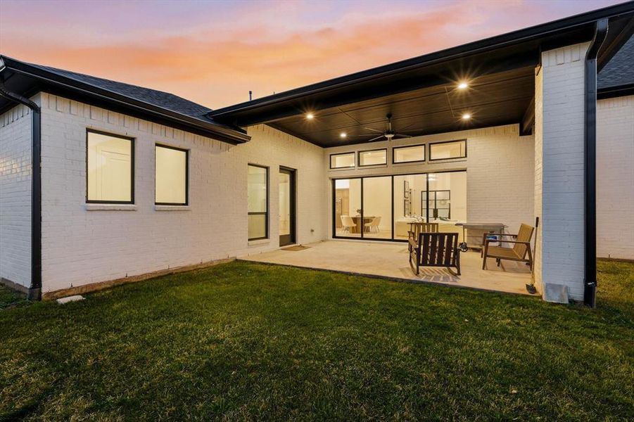 Back of house at dusk with ceiling fan, brick siding, a patio, and a lawn
