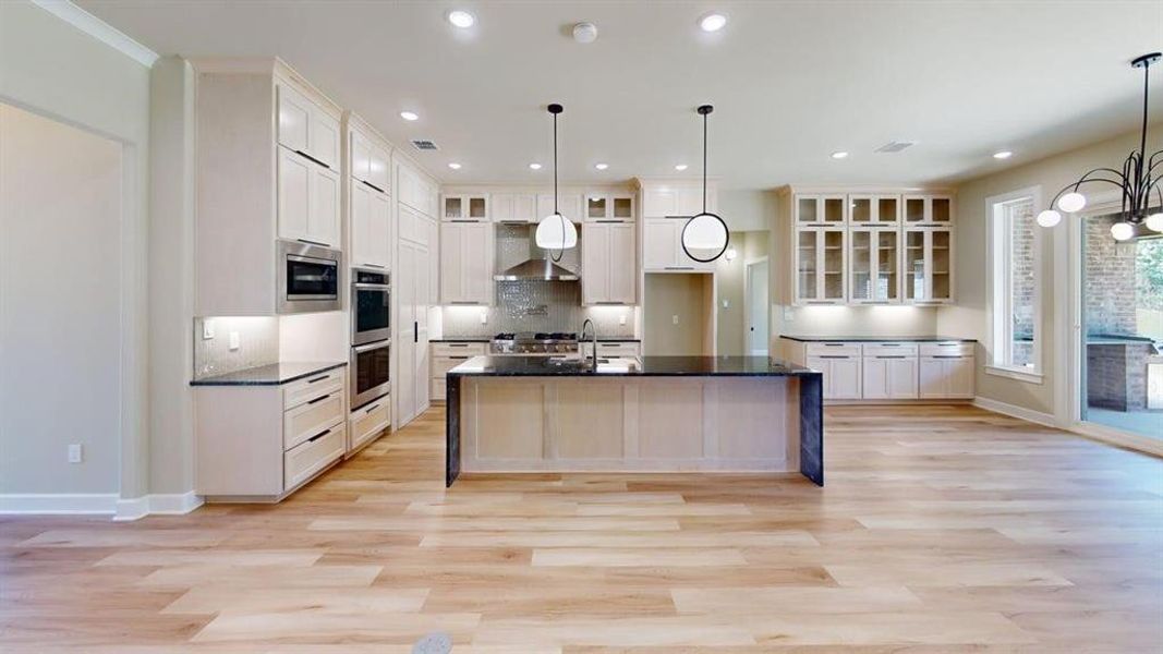 Kitchen with wall chimney exhaust hood, a center island with sink, appliances with stainless steel finishes, glass insert cabinets, and baseboards