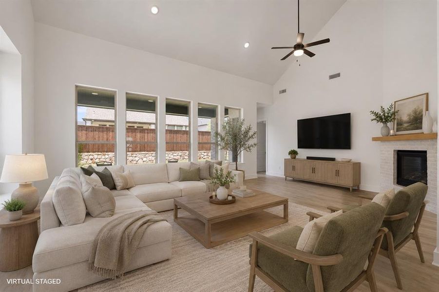 Vaulted ceiling living area featuring wood-finish flooring, a white brick fireplace with a wood mantel, a ceiling fan, recessed lighting, and multiple tall windows Vaulted ceiling living area featuring wood-finish flooring, a white brick fireplace with a wood mantel, a ceiling fan, recessed lighting, and multiple tall windows