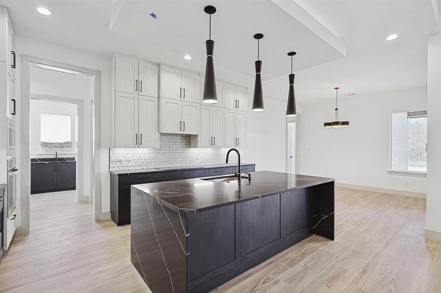 Kitchen with hanging light fixtures, a kitchen island with sink, dark cabinetry, light wood finished floors, and decorative backsplash Kitchen with hanging light fixtures, a kitchen island with sink, dark cabinetry, light wood finished floors, and decorative backsplash