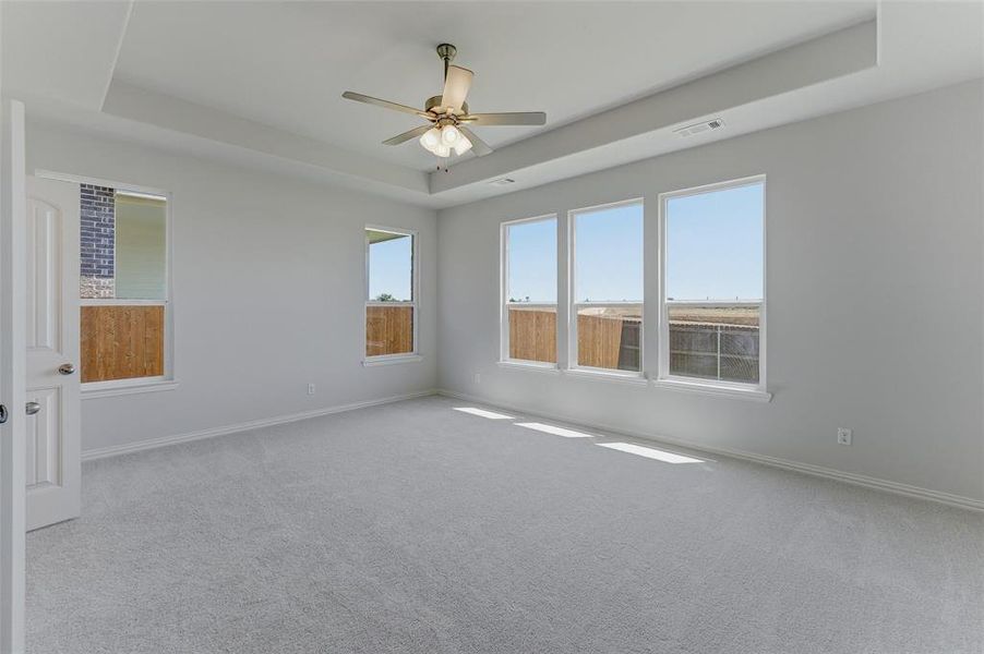 Spacious room featuring light gray carpet, multiple windows, a tray ceiling, and a ceiling fan with integrated lighting