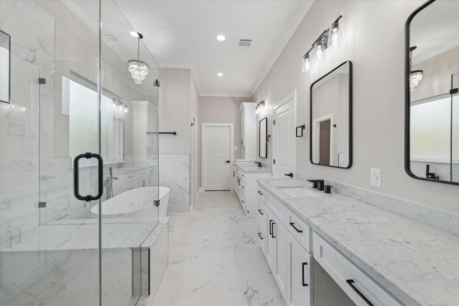 Primary bath with 7ft frameless shower and seat.  Garden soaking tub. Two counters with framed mirrors above and crown molding.  Vanity area on one counter.  Door between counters leads to primary closet.  Door at the opposite end of the room leads to the utility room. Mini chandelier hangs over the tub. Potty room door is just beyond the tub on the left.