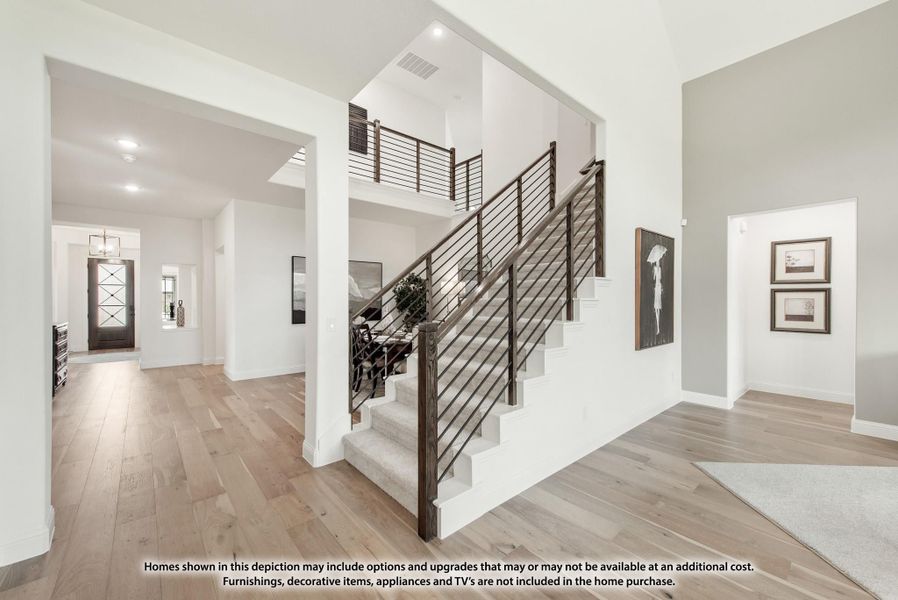 Open foyer with carpeted staircase, dark metal railing, and light hardwood floors leading to front entry door.