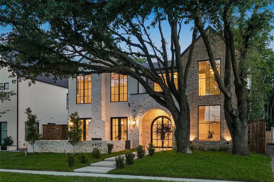 Contemporary facade featuring a prominent arched entry, brick and stone exterior, large black-framed windows, manicured lawn, and mature trees