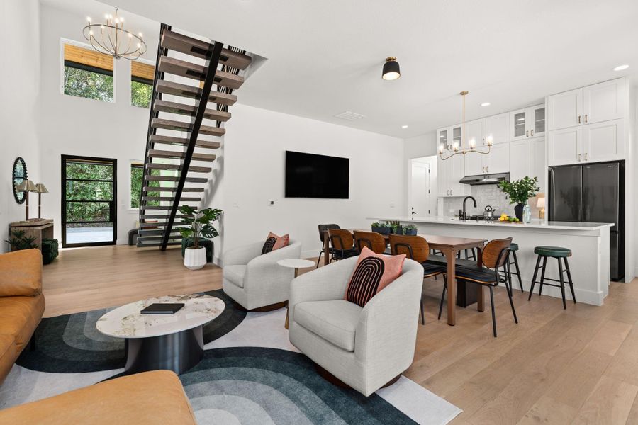 This modern open-concept living space features a stylish kitchen with white cabinets, a large island, and chic lighting. The living area has plenty of room for comfortable seating and a sleek staircase, with abundant natural light and a seamless flow perfect for entertaining.