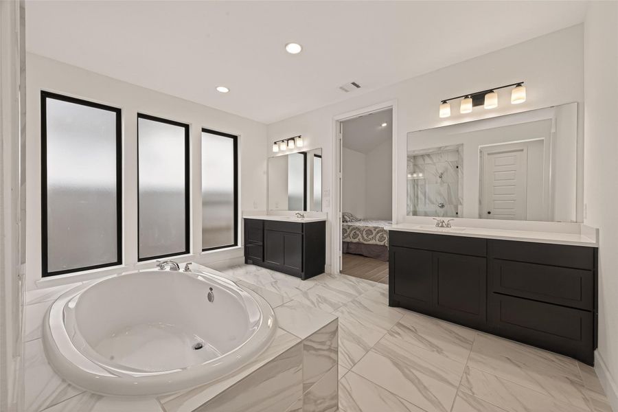 This spacious bathroom features a large soaking tub, dual vanities with dark cabinetry, and frosted windows for privacy. The neutral palette and modern fixtures provide a sleek, inviting atmosphere.