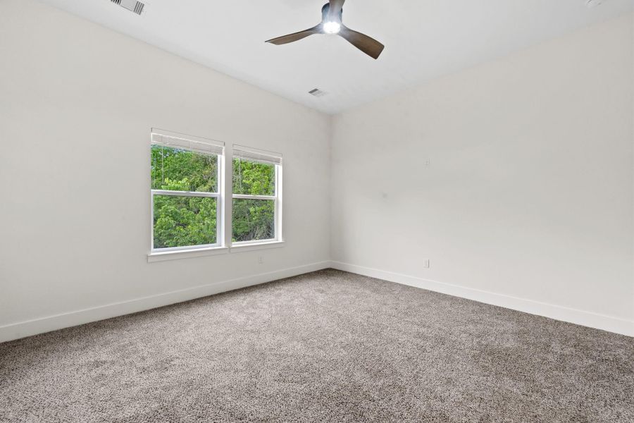 Spacious bedroom with plush carpeting, a modern ceiling fan, and large windows offering serene views of lush greenery.