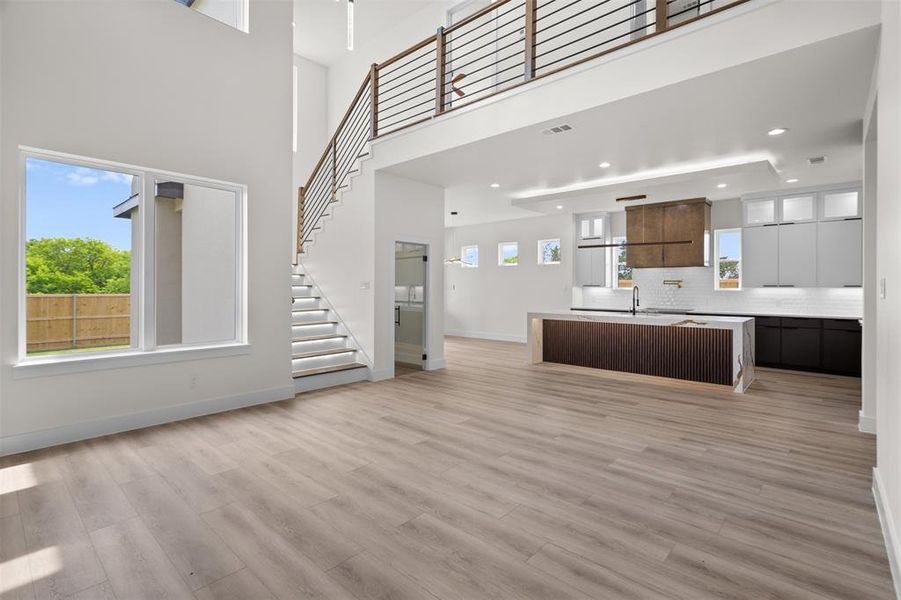 Expansive open-concept living area featuring light-toned flooring, a modern staircase with a dark railing, and a kitchen with white and dark cabinetry Expansive open-concept living area featuring light-toned flooring, a modern staircase with a dark railing, and a kitchen with white and dark cabinetry