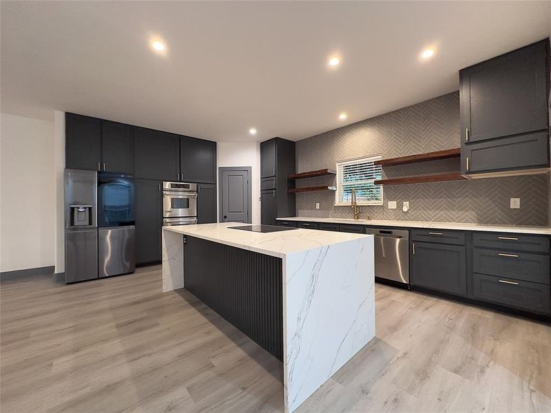 Kitchen with appliances with stainless steel finishes, light stone counters, light hardwood / wood-style floors, backsplash, and an island with sink