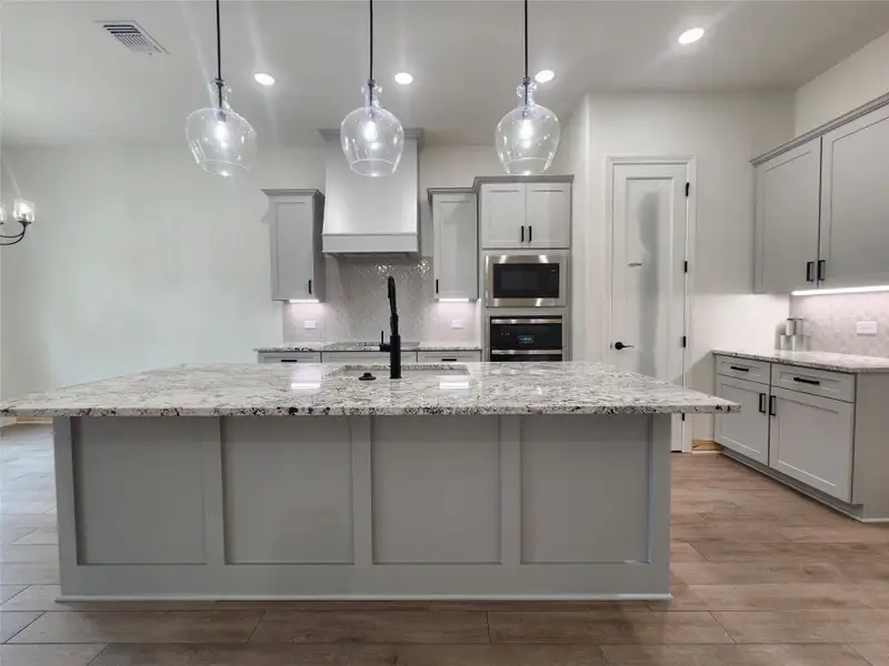 Spacious kitchen featuring a large island with a light-toned countertop, gray shaker-style cabinetry, integrated stainless steel appliances, a geometric tile backsplash, and wood-finish flooring