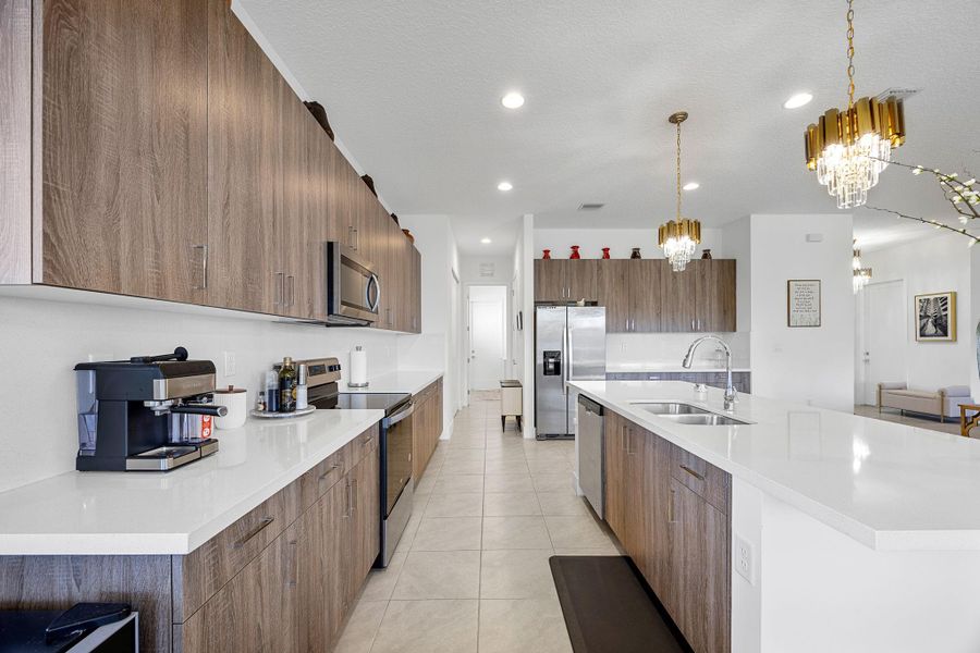 Kitchen featuring modern cabinets, appliances with stainless steel finishes, decorative light fixtures, a center island with sink, and light stone counters
