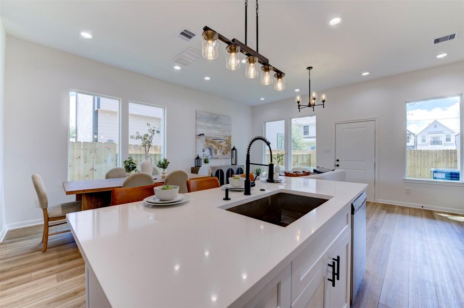 Kitchen island detail with quartz countertop, farmhouse sink, matte black coil-spring faucet, Edison rail pendant, and open view through to dining area and rear windows with yard and street views.