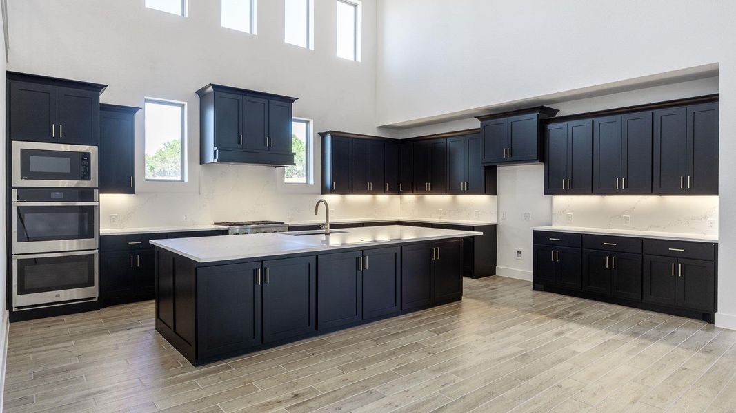 Kitchen with dark cabinets, appliances with stainless steel finishes, an island with sink, a high ceiling, and healthy amount of natural light