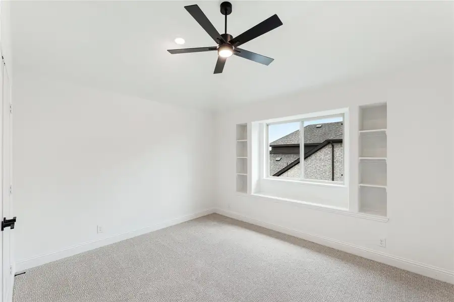 Empty bedroom with carpet, black ceiling fan, and built-in shelves flanking a window