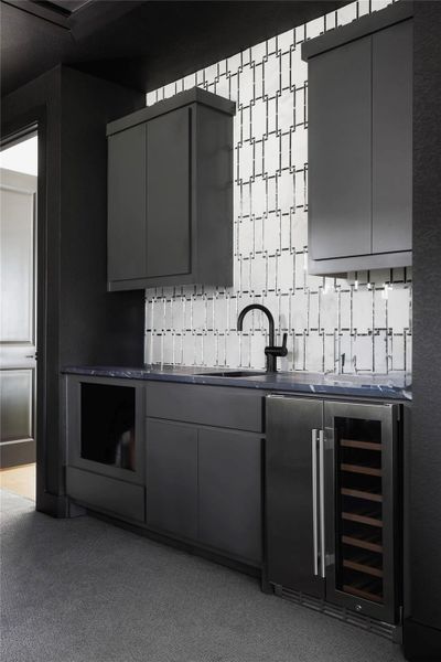 Wet bar featuring dark cabinetry, a patterned tile backsplash, dark countertop with undermount sink, and a stainless steel beverage cooler