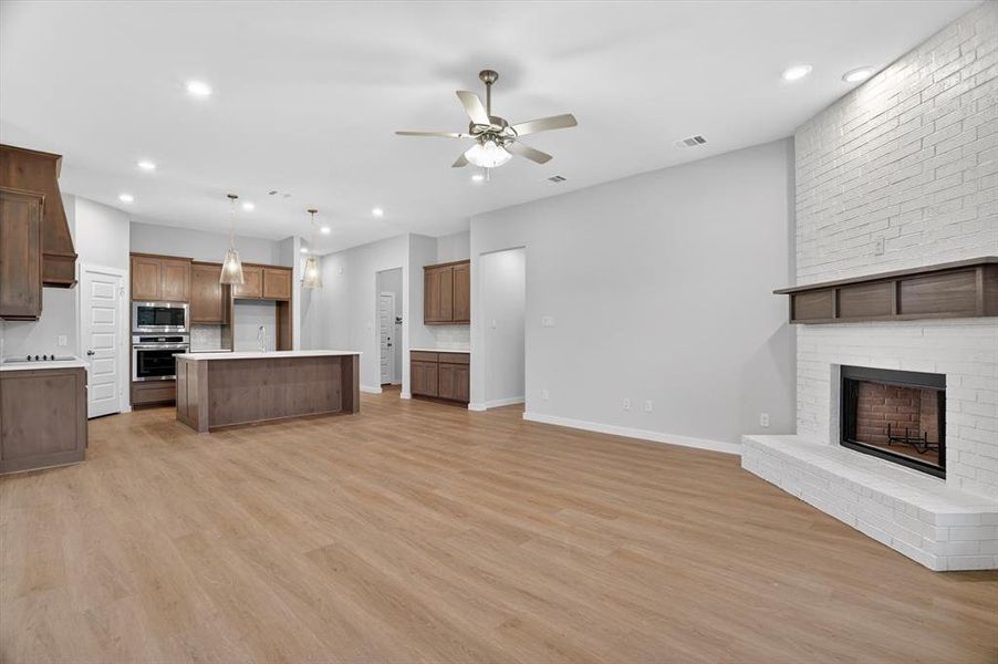Open-concept interior featuring light wood-finish flooring, a white brick fireplace with a wood mantel, a kitchen island with a white countertop, recessed lighting, and a ceiling fan