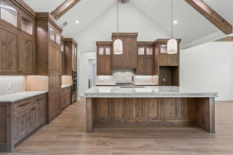 Kitchen featuring brown cabinetry, a large island with sink, pendant lighting, light stone counters, and backsplash Kitchen featuring brown cabinetry, a large island with sink, pendant lighting, light stone counters, and backsplash