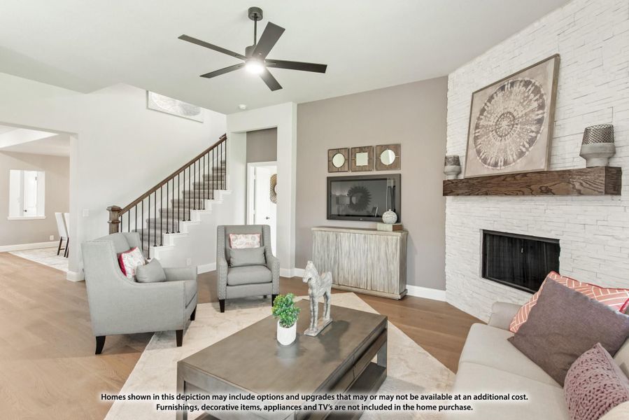 Living room with white stone fireplace, ceiling fan, hardwood floors, and staircase in background
