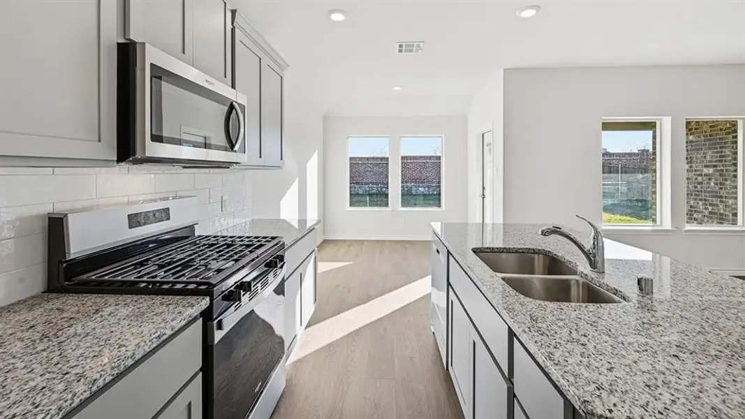 The kitchen features granite countertops, light grey cabinetry, stainless steel appliances including a microwave and gas range, and an island with a double basin sink