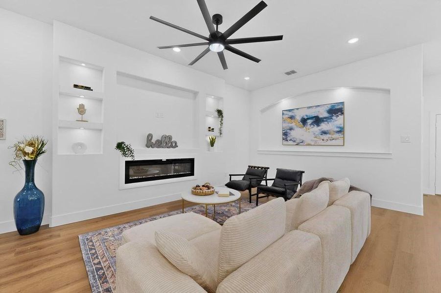 Living area with a glass covered fireplace, light wood finished floors, built in shelves, a ceiling fan, and recessed lighting Living area with a glass covered fireplace, light wood finished floors, built in shelves, a ceiling fan, and recessed lighting