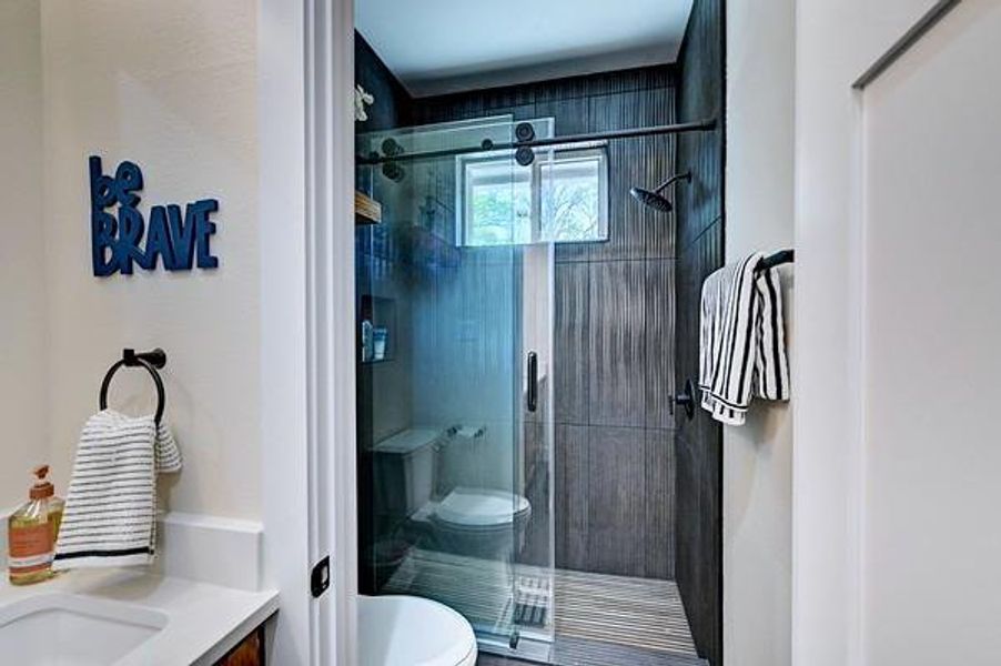 This bathroom features a modern design with a sleek walk-in shower enclosed by glass doors, dark textured wall tiles, and a small window for natural light. A white countertop with a sink is complemented by contemporary fixtures, and the space is accentuated by a motivational wall sign.