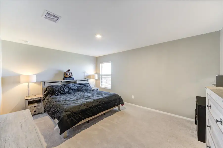 This spacious bedroom features neutral walls, carpeted flooring, and ample natural light from a large window. It includes a modern bed, side tables with lamps, and a dresser, offering a cozy and inviting atmosphere.