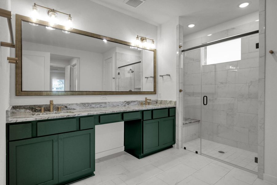 The large vanity has a solid counter top, dual sinks, gold finished faucets, lighting and towel bars plus a large framed mirror. The oversized frameless shower has a bench seat.