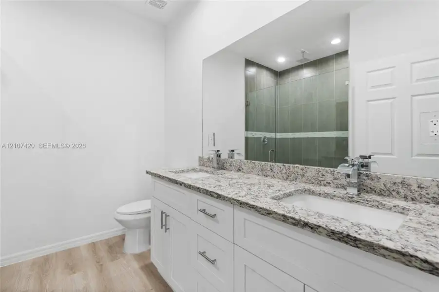 Dual-sink vanity with stone-finish countertop, white cabinetry with silver-tone hardware, wood-finish flooring, and a walk-in shower with tiled surround Dual-sink vanity with stone-finish countertop, white cabinetry with silver-tone hardware, wood-finish flooring, and a walk-in shower with tiled surround