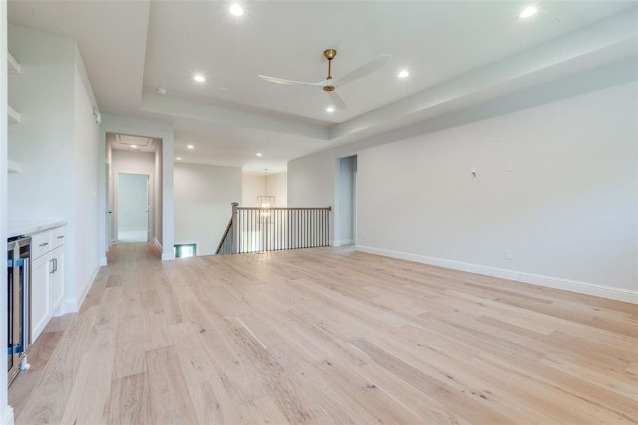 Unfurnished living room featuring light wood-type flooring, a tray ceiling, recessed lighting, a ceiling fan, and beverage cooler Unfurnished living room featuring light wood-type flooring, a tray ceiling, recessed lighting, a ceiling fan, and beverage cooler