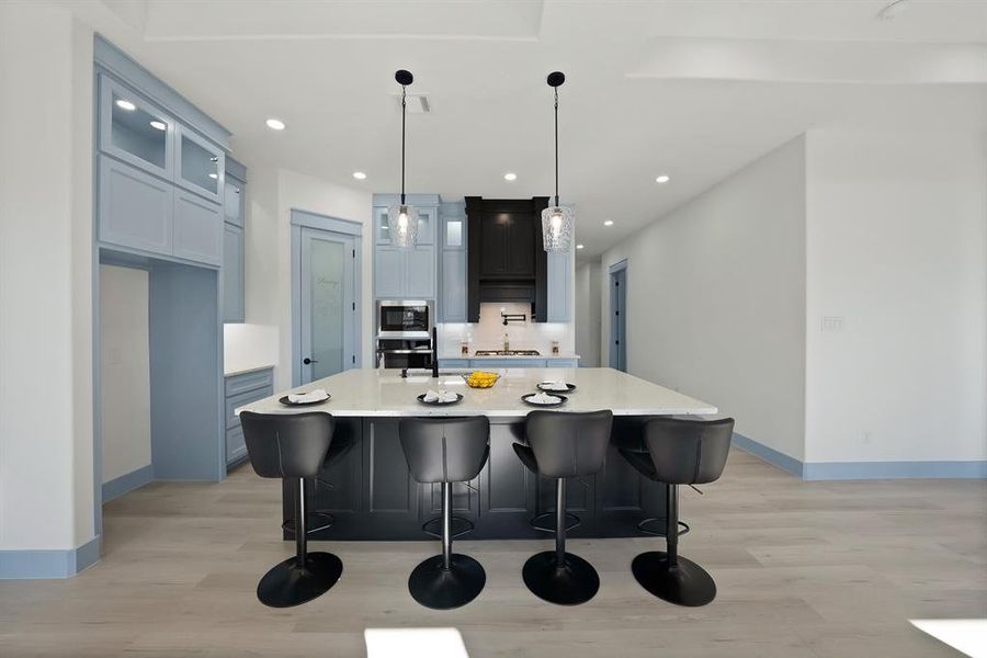 Kitchen with a breakfast bar, hanging light fixtures, a center island with sink, light wood-style flooring, and glass insert cabinets