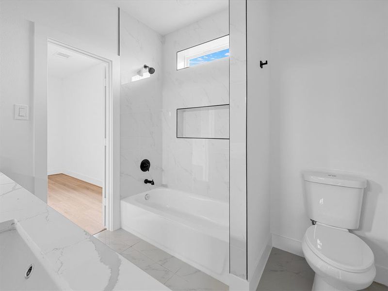 This bathroom features a sleek, modern design with white marble tiles, a bathtub with black fixtures, a toilet, and a window for natural light. The entrance opens to a room with light wood flooring.