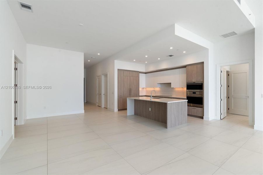Open-concept kitchen featuring two-toned cabinetry, stone-finish countertops, a central island with an integrated sink, built-in stainless steel appliances, and large-format tile flooring