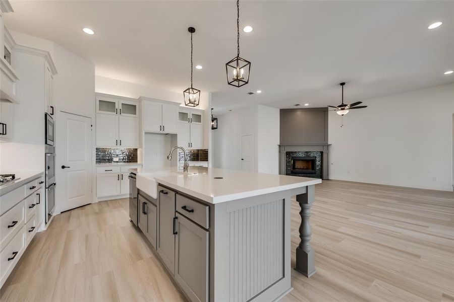 Kitchen with a stone fireplace, light hardwood / wood-style floors, tasteful backsplash, an island with sink, and hanging light fixtures