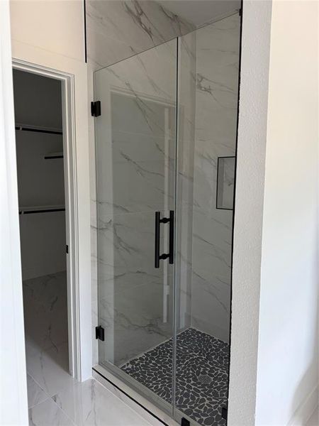 This photo shows a modern walk-in shower with marble-like tile walls, a clear glass door with black hardware, and a stylish pebble tile floor.