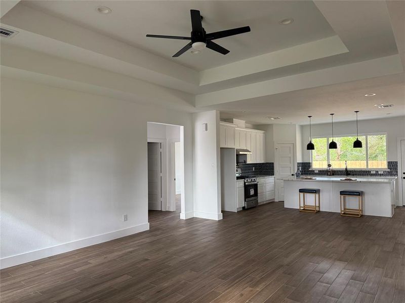 This photo features a spacious, open-concept living area with a modern kitchen, including a central island with bar seating, stainless steel appliances, and stylish pendant lighting. The room is bright with ample natural light, showcasing wood flooring and high ceilings with recessed lighting and a ceiling fan.