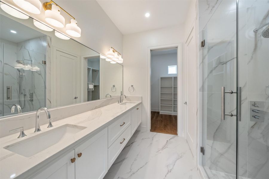 Primary bathroom features a modern design with a double vanity, sleek marble countertops, and a large mirror. It includes a glass-enclosed shower and ample storage space, leading to a walk-in closet. The space is bright, with elegant lighting and a clean, polished look.
