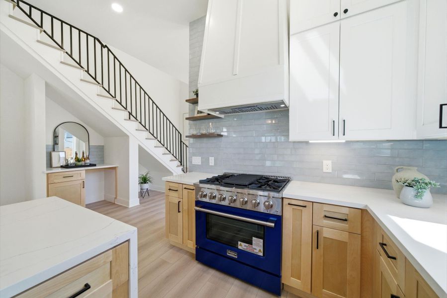 This modern kitchen features sleek white cabinets and light wood accents, complemented by a striking blue range. The glossy subway tile backsplash adds a touch of elegance, while an open stairway with a black railing enhances the space's contemporary feel.