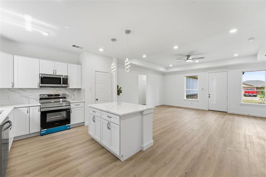 Modern kitchen and living area featuring a central island with pendant lighting, stainless steel appliances, white cabinetry with contemporary hardware, and wood-finish flooring