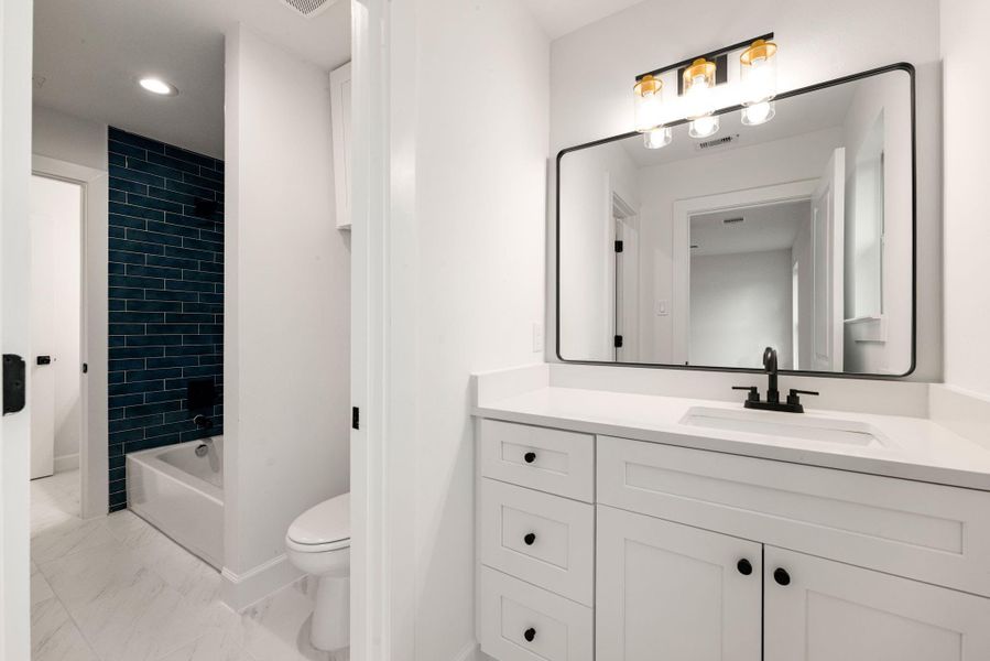 Modern bathroom featuring a sleek white vanity with quartz countertop, black hardware, and a large framed mirror with contemporary lighting. The space is complemented by stylish tile flooring and a tub/shower combo with striking dark tile accent for a bold, modern touch.