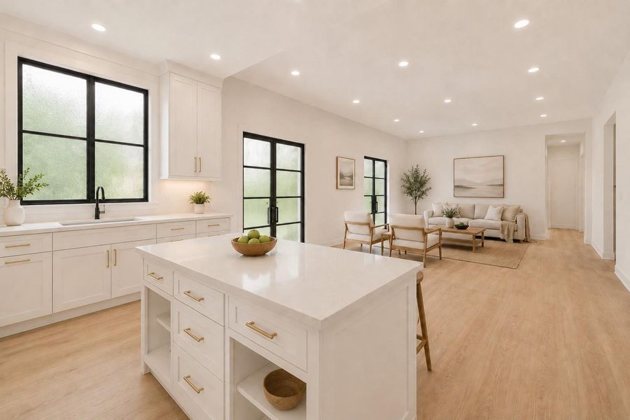 Open-concept kitchen and living area featuring a white kitchen island with built-in storage, white cabinetry with brass hardware, a black-framed window above the sink, and wood-finish flooring throughout