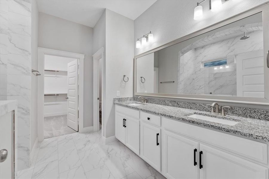 This is a modern bathroom featuring a double vanity with granite countertops and white cabinetry, large mirror, marble-style flooring and wall tiles, and sleek fixtures.