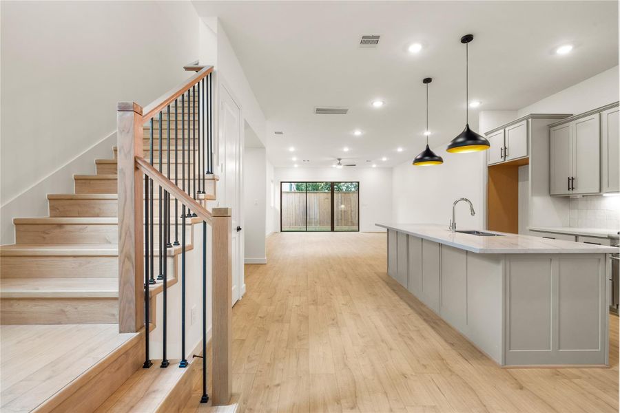 Grand main-level floor plan — expansive quartz kitchen island, matte black dome pendant lights, custom wood-and-iron staircase, and floor-to-ceiling glass patio doors.