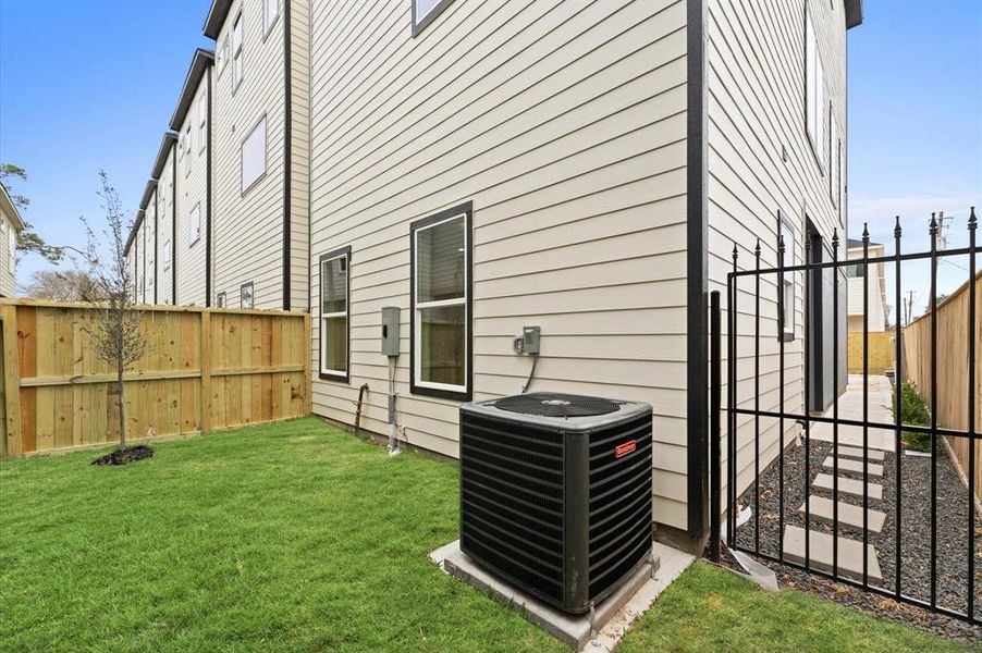 This photo shows the side of a modern townhouse with beige siding, featuring a small fenced backyard with grass and a young tree. There's an air conditioning unit and a narrow pathway with stepping stones leading to a gate.