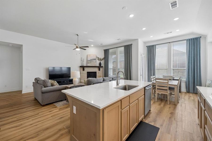 Open-concept living space featuring wood-finish flooring, a kitchen island with an undermount sink, recessed ceiling lighting, a stone fireplace, and multiple large windows with blinds
