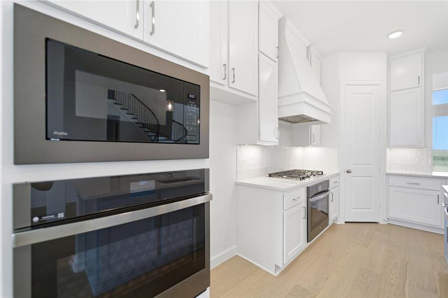 Kitchen featuring white shaker cabinetry, light wood-finish flooring, a built-in stainless steel microwave, a built-in stainless steel oven, and a gas cooktop with a custom range hood