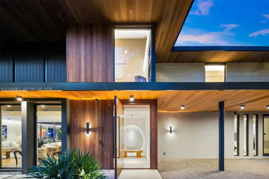 Contemporary facade featuring extensive wood-paneled cladding, floor-to-ceiling glass windows, recessed ceiling lighting, a wide pivot entry door, and covered stone paver driveway