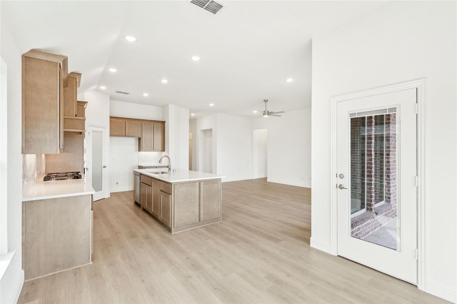 Open-concept kitchen with center island, tan cabinets, light wood floors, and adjacent living area with ceiling fan. Open-concept kitchen with center island, tan cabinets, light wood floors, and adjacent living area with ceiling fan.
