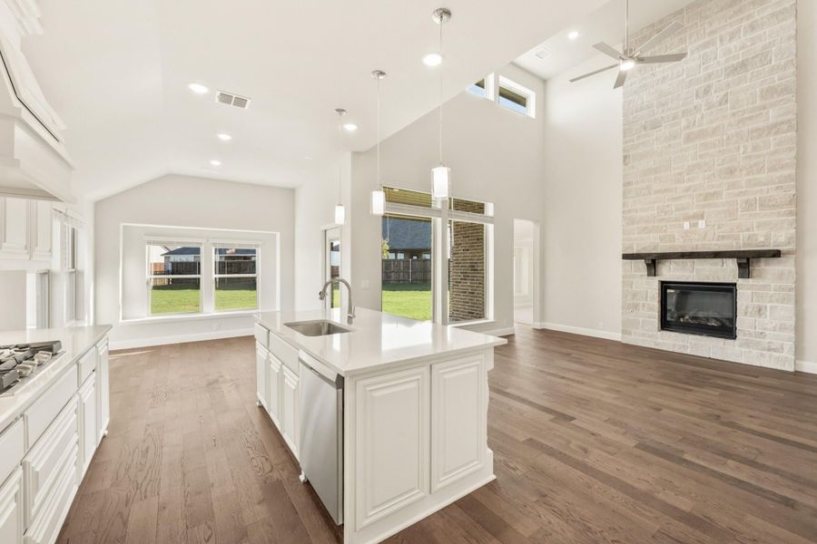 Open-concept kitchen with white island, hardwood floors, and stone fireplace in adjoining living area