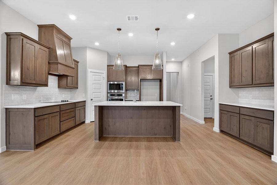 Spacious kitchen featuring an expansive center island, wood-finish flooring, custom wood cabinetry, stainless steel appliances, and pendant lighting